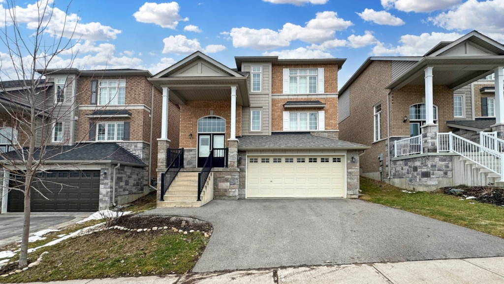 A two-story brick house with a double garage and central front entrance beckons potential buyers. Steps lead up to the entrance, surrounded by similarly designed homes. A real estate agent would appreciate the cloudy sky and patches of grass and snow enhancing its charm.