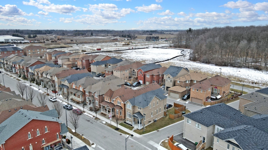 Aerial view of a suburban neighborhood, perfect for a real estate agent interested in buying and selling properties. Rows of similar-style houses boast varying colored roofs. Streets lined with bare trees and patches of snow surround the homes, adjacent to wooded areas and open land.