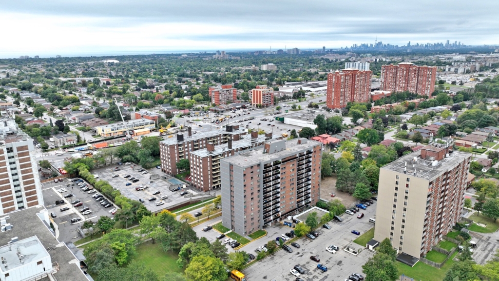 Aerial view of a suburban area with several mid-rise apartment buildings, parking lots, and green spaces. The city skyline is visible in the distance under a cloudy sky.